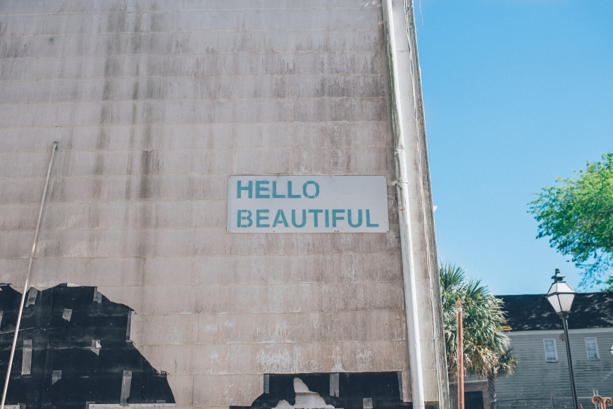 A photo of the side of a large building with a plain, pale grey wall and little else on it aside from a sign that says 'Hello beautiful' in blue. In the background to the right hand side is the street where you can see a roof, a streetlight, some trees and the blue sky.