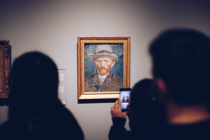 A photo of a self portrait by Van Gogh hanging in a gallery, with silhouettes of people in the foreground with one of them holding up a smartphone with the camera open.