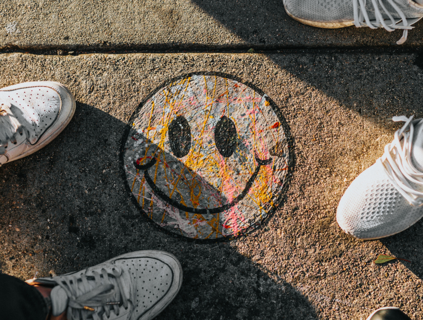 A multi-colored smiley face drawn with chalk on a pavement ground, photographed from above. Two pairs of trainers can be seen next to the drawing, implying two people standing around it.