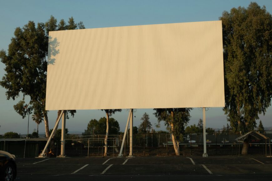 A photo of a blank white billboard against blue sky, with trees in the background.